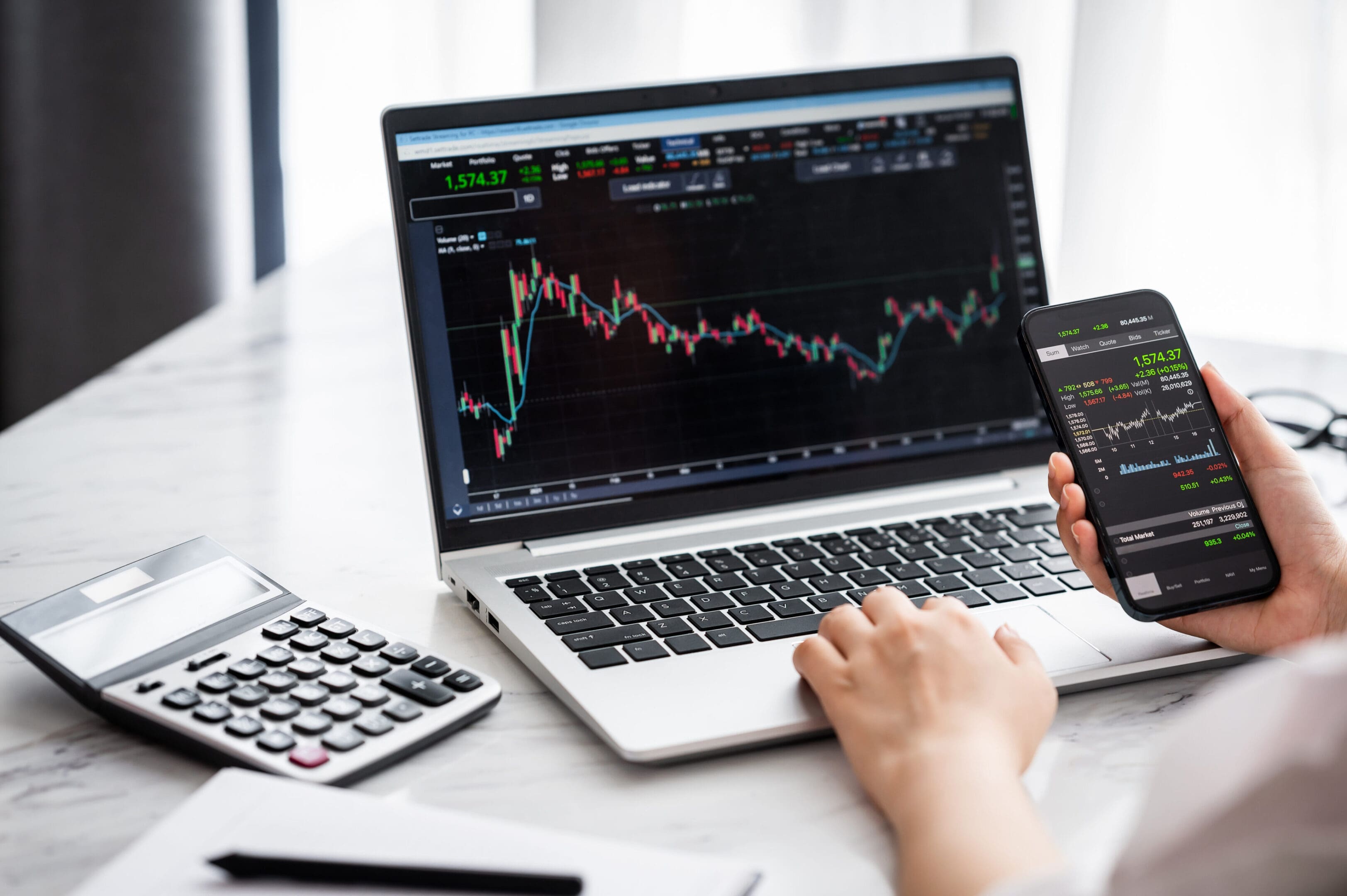 Hand holding smartphone with stock market data and using laptop display graph and chart for analyze and check before trading stocks online. Depicts TradingView financial market chart.
